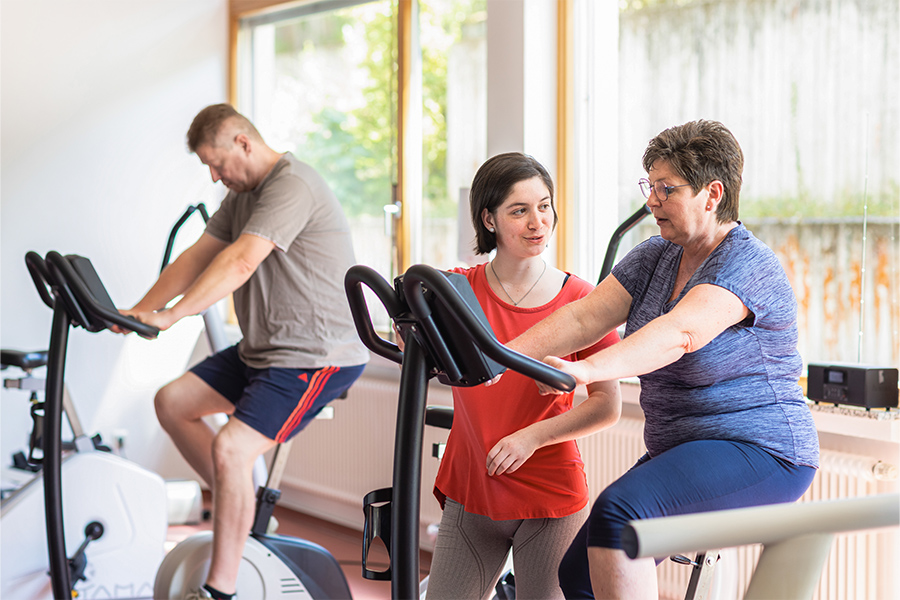 Zwei Patienten trainieren im Fitnessstudio. Eine Therapeutin unterstützt eine Patientin beim Training auf dem Ergometer.